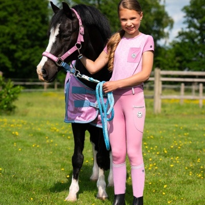 Criança com roupa rosa a segurar pónei preto e branco com arreios rosa num campo verde com flores amarelas