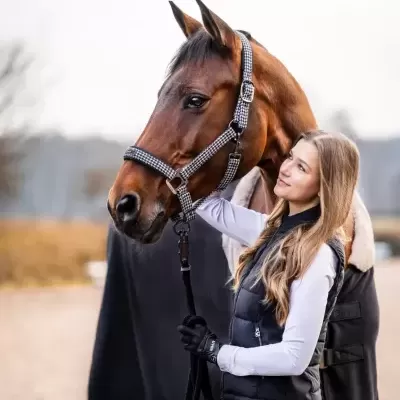 Jovem com cavalo castanho usando cabresto preto e branco e roupa de equitação preta.