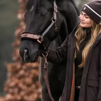 Cavalo preto com cabresto cinzento e mulher com casaco e gorro castanhos ao ar livre
