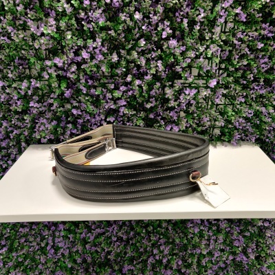 Alça de bag preta com fivela metálica sobre superfície branca e fundo de flores lilases.
