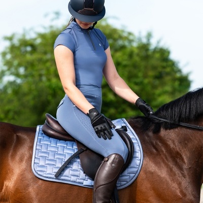 Mulher com roupa de equitação azul clara e capacete preto montada num cavalo castanho com manta azul clara acolchoada