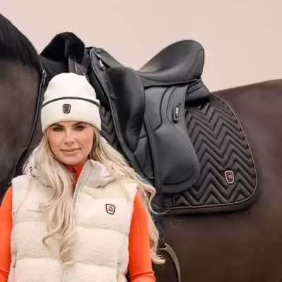 Mulher com colete e gorro bege em tecido felpudo e cavalo castanho com sela preta.