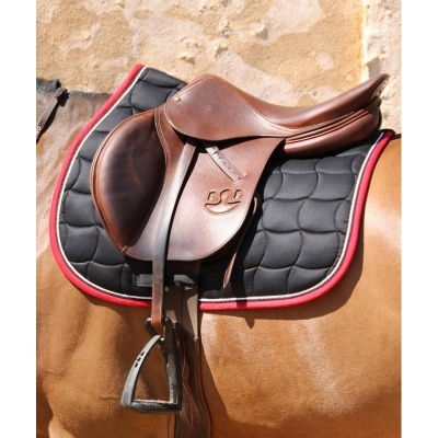 Sela de couro castanho sobre manta preta com borda vermelha, montada em cavalo castanho