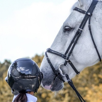 Pessoa com capacete preto e camisa branca olhando para cavalo branco com rédeas pretas ao ar livre.