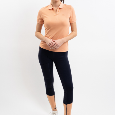 Mulher de pé com polo pêssego e calças justas negras e bege, calçado branco, fundo branco.