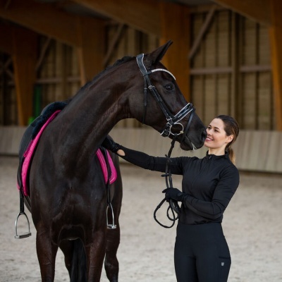 Cavalo castanho escuro com sela preta e manta rosa com mulher vestida de preto em picadeiro
