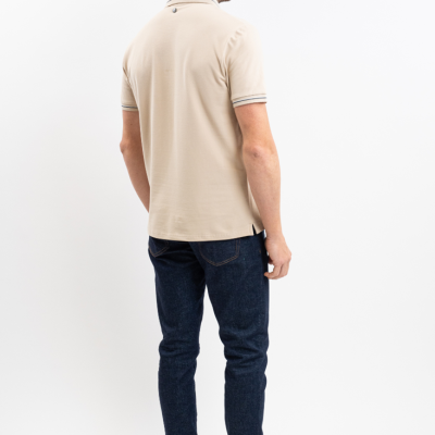 Homem de costas com polo bege e jeans azul escuro num fundo branco