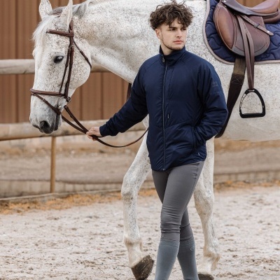 Homem com casaco azul e calças de equitação cinzentas a conduzir cavalo branco com sela castanha.
