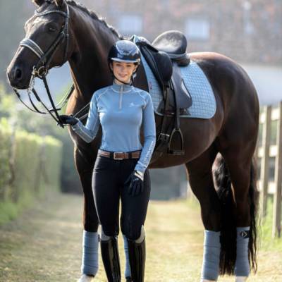 Jovem cavaleira com roupa de equitação azul e preta com cavalo castanho e equipamento azul claro ao ar livre numa área gramada.