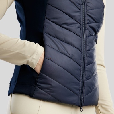 Colete acolchoado azul escuro com fecho, camisa bege e calças bege com detalhe castanho.