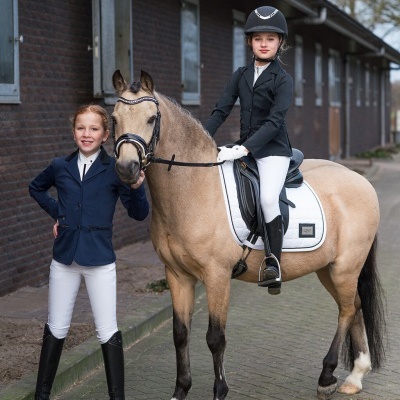 Duas meninas com roupa de equitação e cavalo creme com equipamento branco e preto.