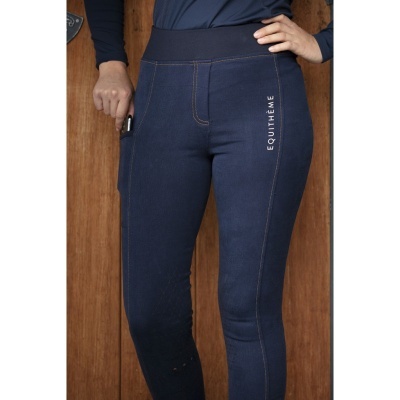 Calças justas azuis escuras com costuras castanhas e texto EQUITHÈME na lateral, pessoa com mão no bolso, fundo madeira