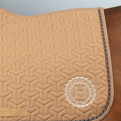 Tapete para sela bege com padrão geométrico e bordado branco 'H' sobre cavalo castanho