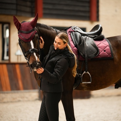 Mulher com casaco preto junto a cavalo com sela e capa bordô para as orelhas
