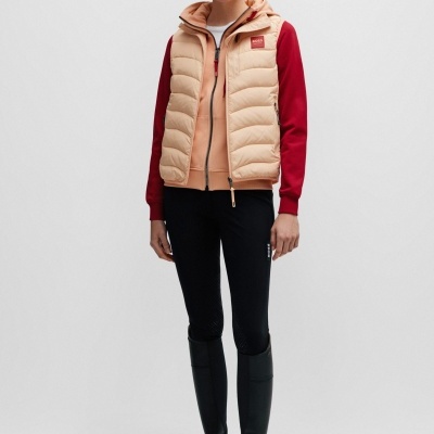 Mulher com colete bege acolchoado, casaco vermelho, leggings pretas, botas pretas e boné cor de pêssego com texto BORSE SPORTSWEAR