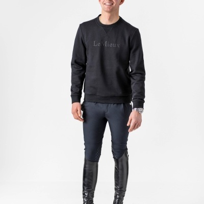 Homem jovem com sweat preto Le Mieux, calças justas pretas e botas de montar pretas em fundo branco