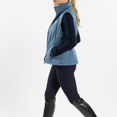 Mulher com colete azul, camisola e leggings pretas, botas pretas altas, fundo branco