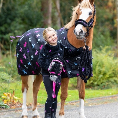 Menina com roupa preta decorada com pezinhos coloridos, segurando cavalinho de pau, ao lado de cavalo com manta e cabresto combinando.