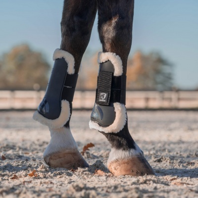 Botas protetoras para pernas de cavalo em pelúcia branca e preto, em solo arenoso ao ar livre