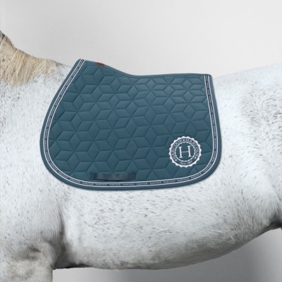 Tapete de sela azul com padrão geométrico acolchoado e logótipo Harcour sobre cavalo cinza claro