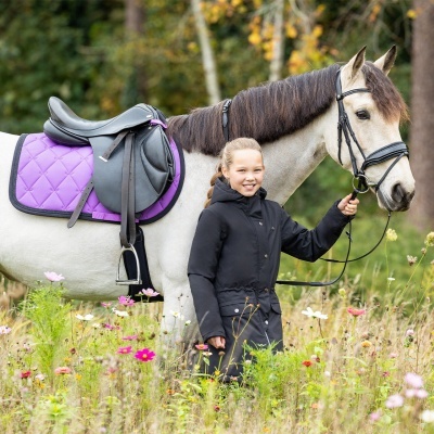 Menina com casaco preto ao lado de cavalo branco com sela preta e manta roxa acolchoada no campo florescente