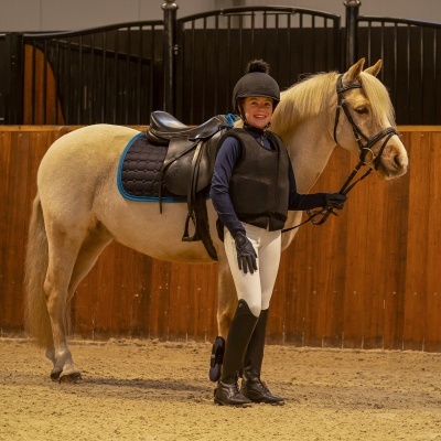 Criança com roupa de equitação ao lado de cavalo claro em picadeiro