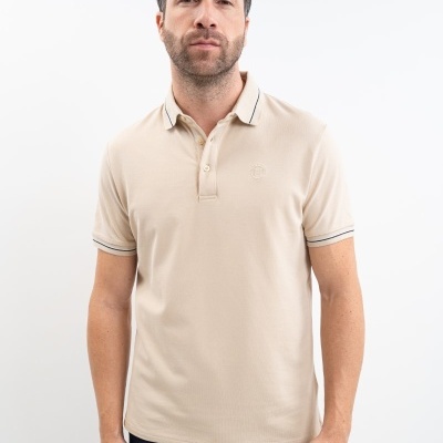 Homem a usar polo bege com riscas nas mangas e gola e jeans escuro em fundo branco