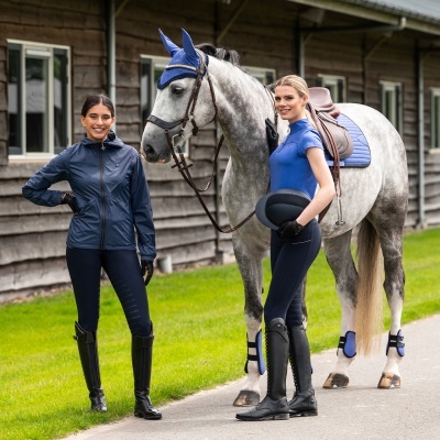 Mulheres com roupa de equitação azul e preta ao lado de um cavalo cinza com acessórios azuis em estábulo