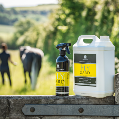 Dois frascos do produto FLY GARD para cavalos sobre madeira com cavalo e pessoa desfocados ao fundo