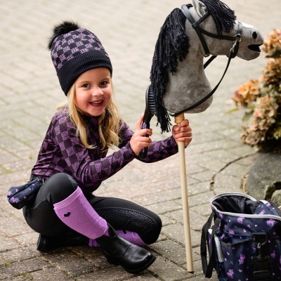 Menina a segurar e escovar cavalo de pau cinzento com crina preta, vestida com roupa preta e violeta, junto a mochila preta com padrão violeta.