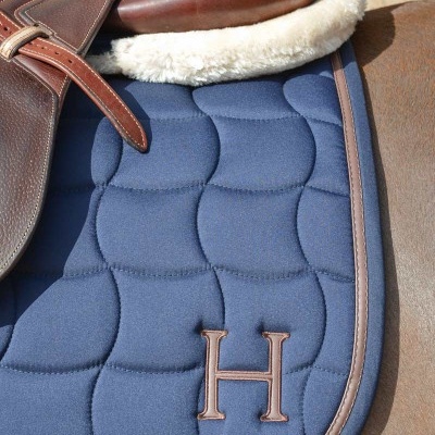 Sela de equitação castanha com almofada acolchoada azul com borda castanha e letra H bordada.