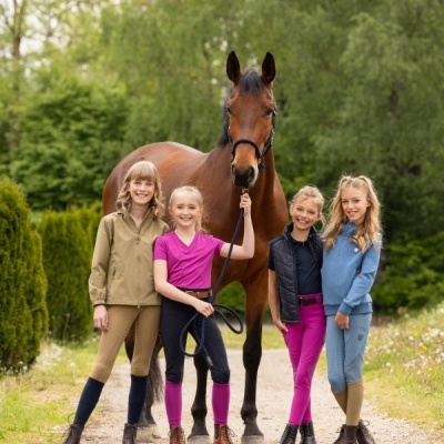 Quatro crianças vestindo roupa de equitação colorida ao lado de cavalo castanho num caminho de terra