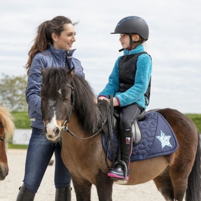 Menina de capacete a montar pónie castanho com tapete azul com estrela, mulher ao lado e pónie castanho claro à esquerda
