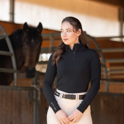 Mulher com roupa de equitação preta e clara num estábulo com cavalo ao fundo