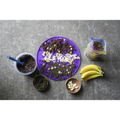 Prato roxo com sementes e pedaços de banana junto a utensílios e ingredientes sobre fundo cinzento.