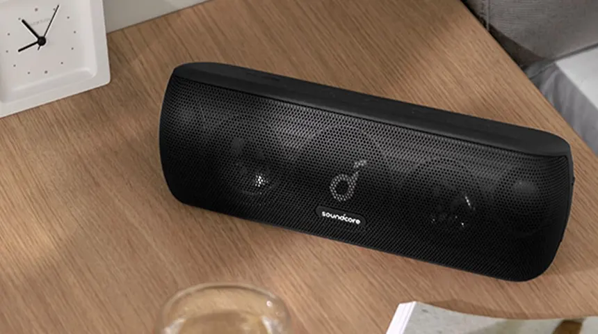 Coluna portátil preta Soundcore sobre mesa de madeira com relógio branco próximo