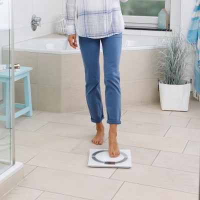 Pessoa usando balança digital branca em casa de banho com planta e móveis azuis