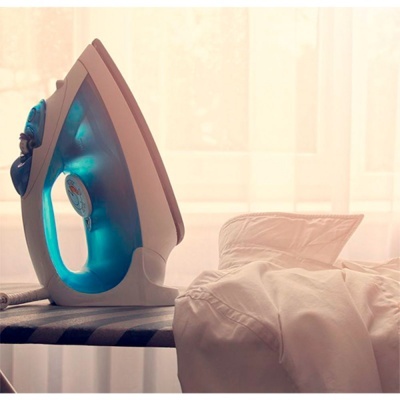 Ferro de engomar azul e branco sobre tábua de engomar com camisa branca e cortinas ao fundo