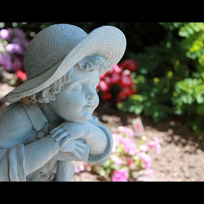 Estátua de criança com chapéu no jardim, rodeada de flores e plantas.