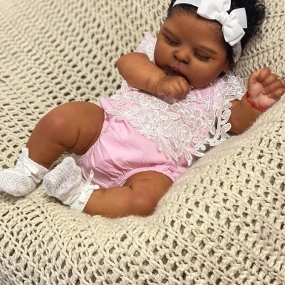 Boneca bebé realista vestida com roupa rosa e renda branca, fita branca na cabeça, deitada numa manta crochê bege.