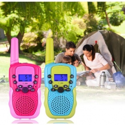 Dois walkie-talkies coloridos com ecrãs digitais, família sentada ao fundo junto a uma tenda no parque