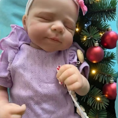 Boneca bebé realista a dormir com roupa lilás, faixa na cabeça e chupeta, junto a decoração de Natal