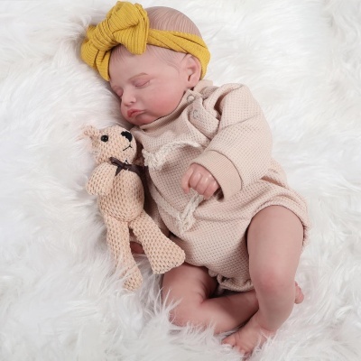 Bebé a dormir com roupa rosa claro e faixa amarela, deitado numa manta branca segurando ursinho de croché.