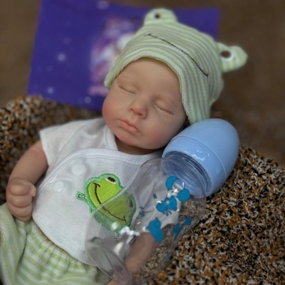 Boneca bebé realista com roupa de rã, deitada com mamadeira azul ao lado