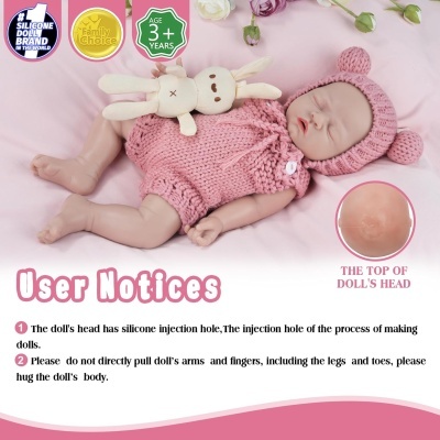 Boneca de silicone realista vestida com fato cor-de-rosa e chapéu com orelhas deitada sobre tecido cor-de-rosa com peluche coelho creme e textos informativos