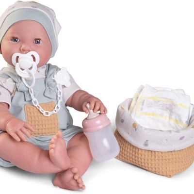 Boneca bebé realista com roupa cinzento-azulada e bege com biberão e chucha, ao lado cesto com fraldas