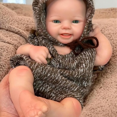 Boneca bebé realista com roupa de malha castanha e branca com capuz, segurada por uma mão sobre tecido castanho.