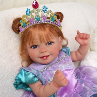 Boneca de bebé com vestido lilás e coroa colorida sentada numa almofada branca