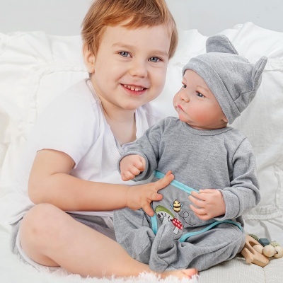 Menino com boneco bebé vestido de cinzento sentado em cama com almofadas brancas