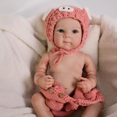 Boneca bebê realista com gorro e saia rosa de malha sentada sobre tecido branco felpudo.